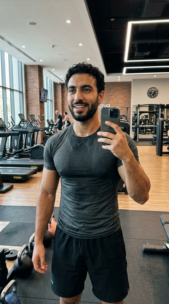صورة سيلفي في صالة رياضية لمدرّب لياقة عربي وسط أجهزة التمرين — شخصية مُولَّدة بالكامل بالذكاء الاصطناعي في عقل بنسبة عمودية 9:16